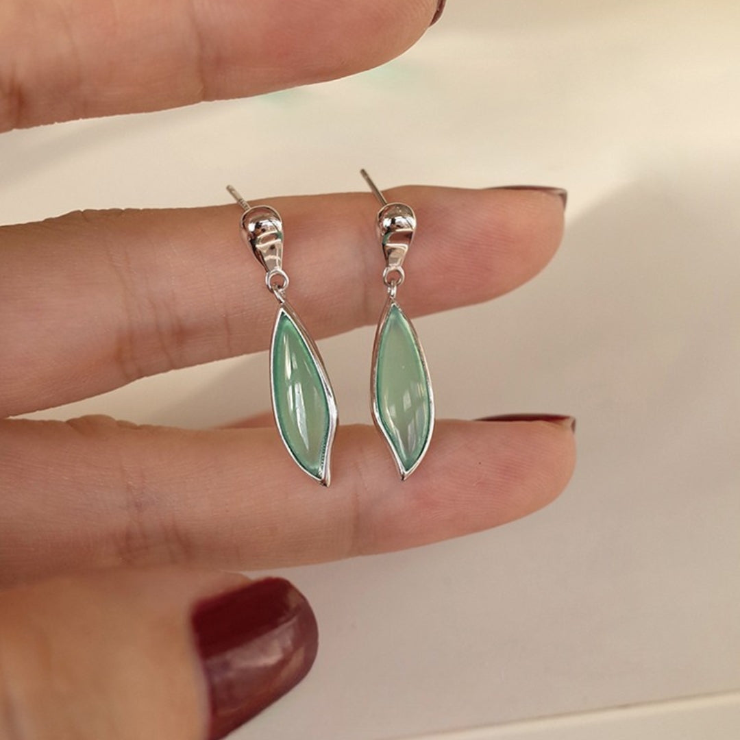 Green leaf earrings with silver hooks held between fingers against a neutral background