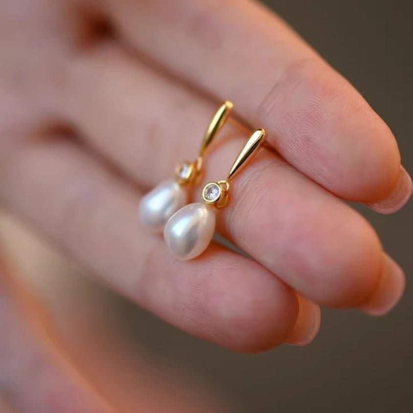 Close-up view of a pair of baroque pearl drop earrings with a geometric gold bar design and zirconia accent held in fingers.