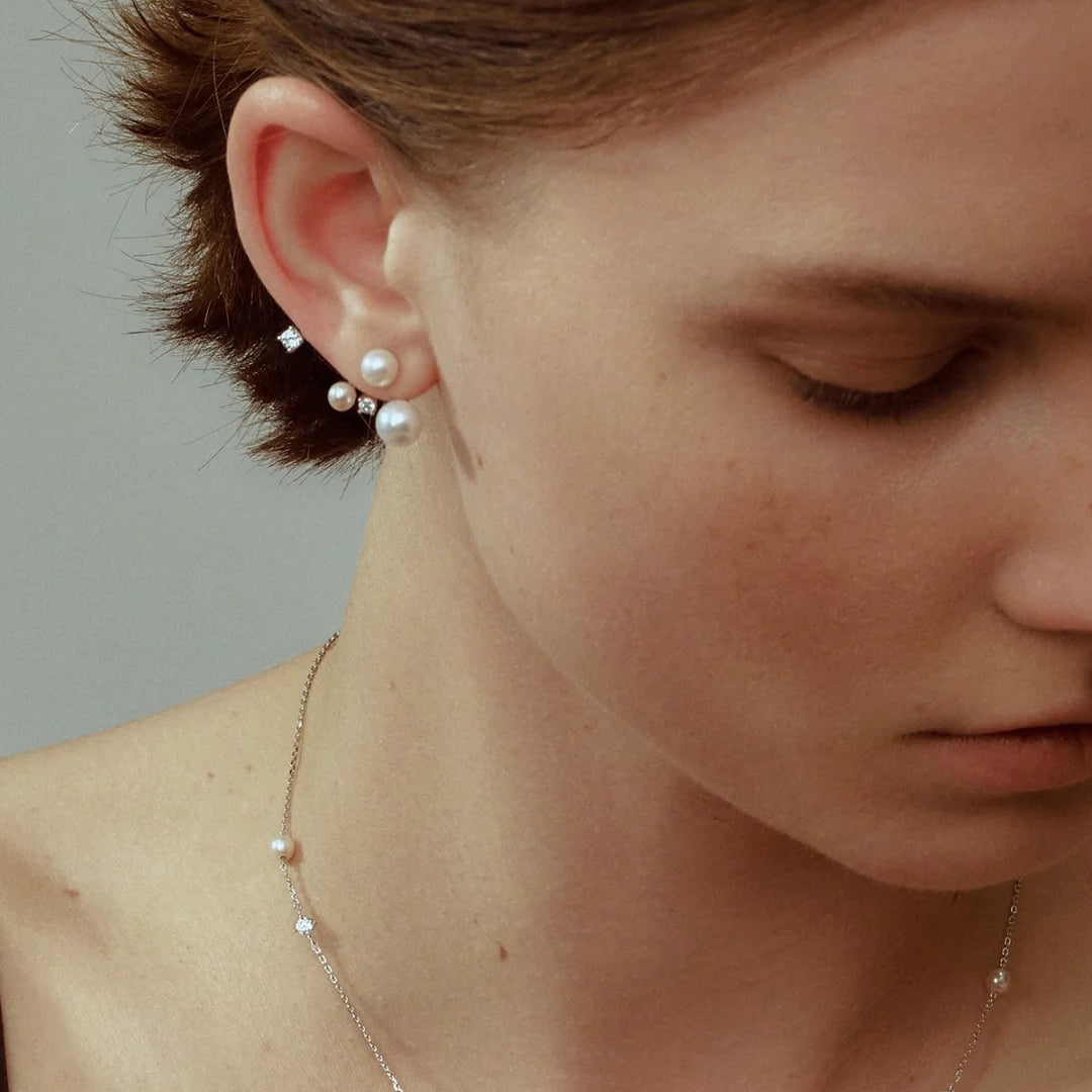 Close-up of a person wearing diamond and pearl earrings and a pearl necklace against a neutral background